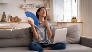 Eine Frau sitzt mit einem Ventilator und einem Laptop auf einem Sofa